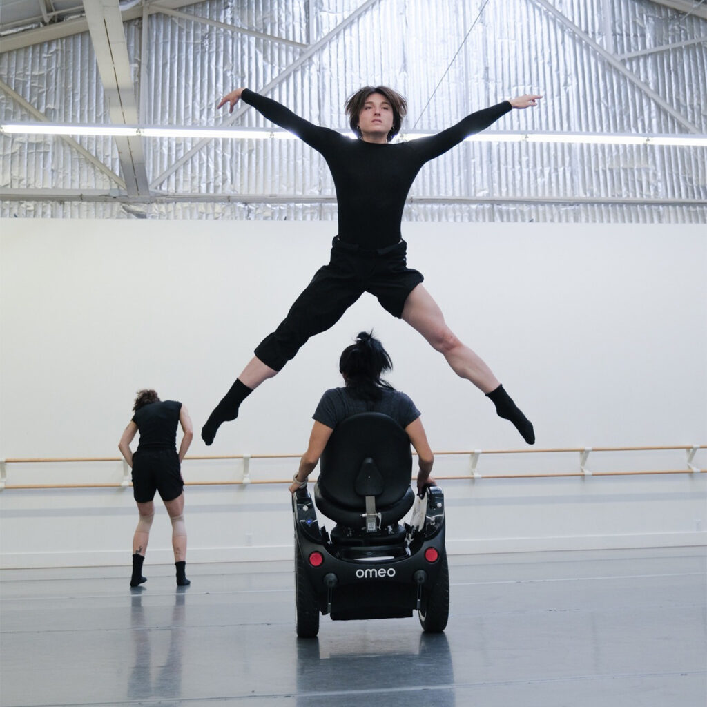 While dancing, a person leaps in the air. Another person sitting in an Omeo moves through their extended legs