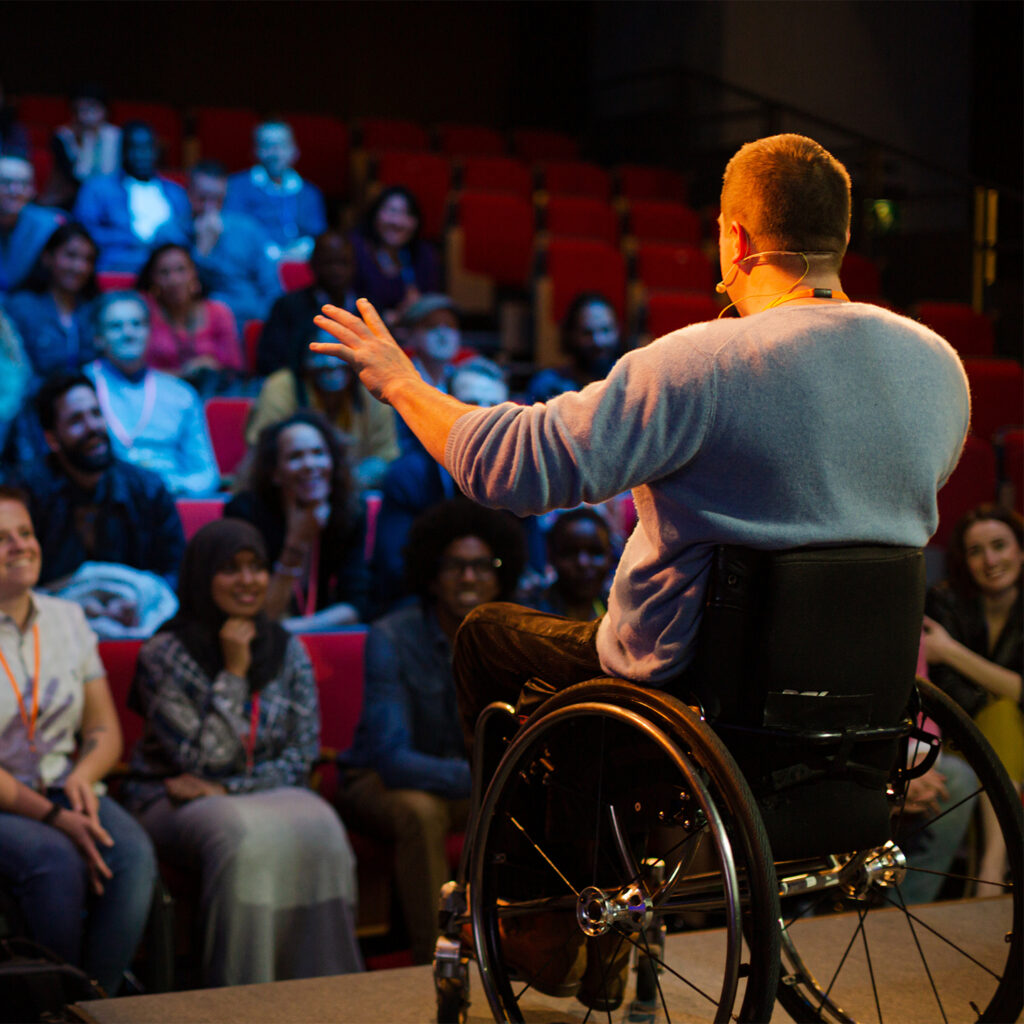 A man in a wheelchair speaks to a crowd. The man is on a raised stage