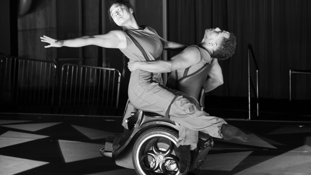 Two dancers perform a contemporary piece, with one seated on an Omeo hands-free mobility device and the other extending gracefully while being supported. The black-and-white image captures the power and fluidity of inclusive movement and assistive technology in performance.
