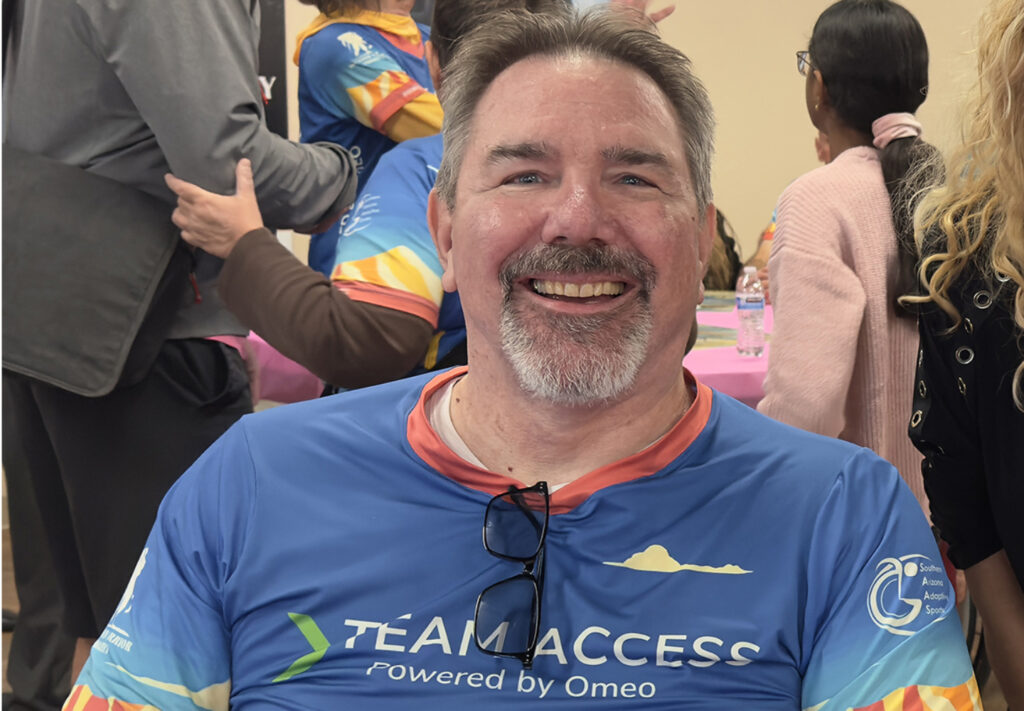 A smiling man wearing a colorful 'Team Access – Powered by Omeo' jersey sits indoors at a community event. People in similar jerseys and casual attire gather around tables in the background.