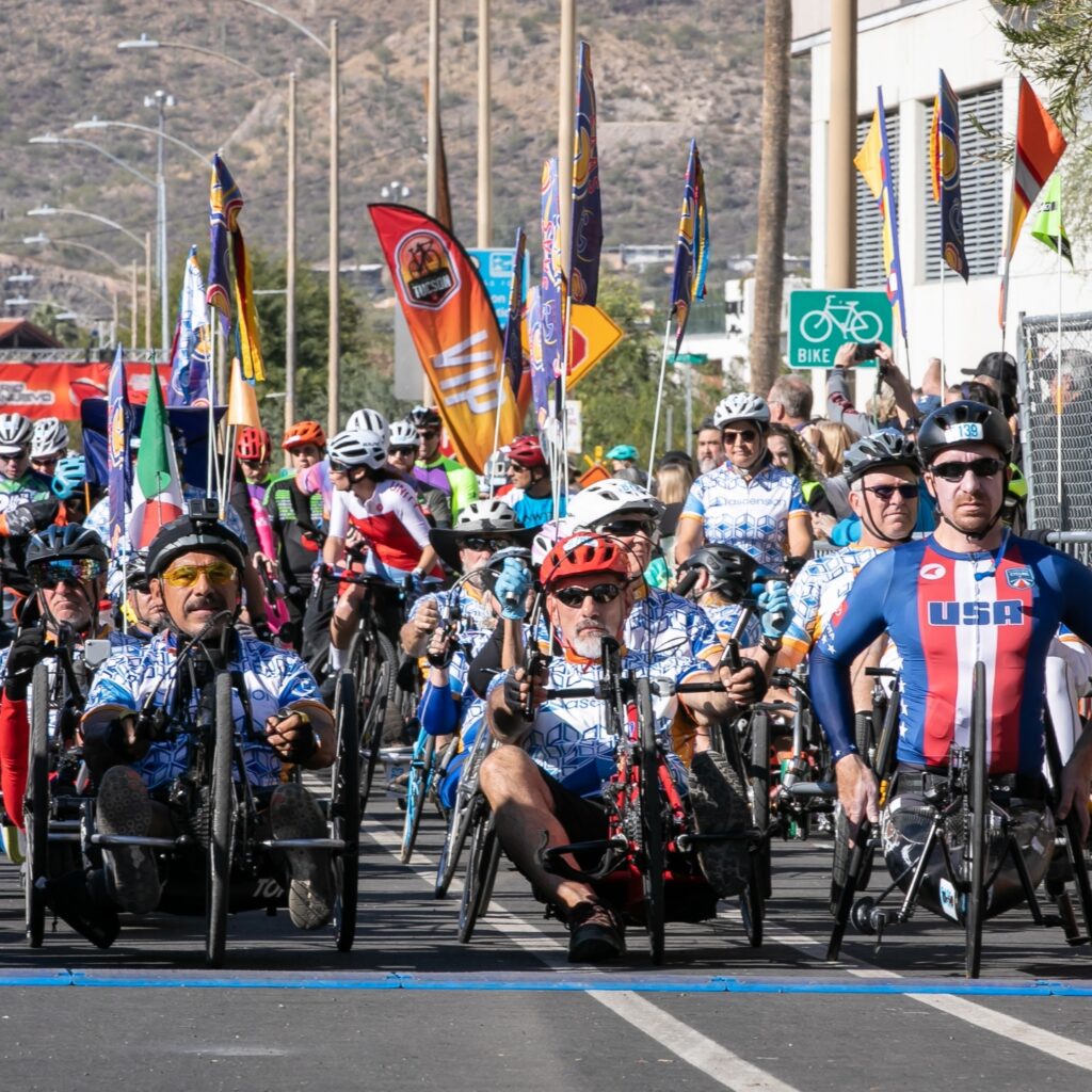 Dozens of adaptive cyclists gather at the El Tour de Tucson starting line, surrounded by event flags and spectators, preparing to begin the race.