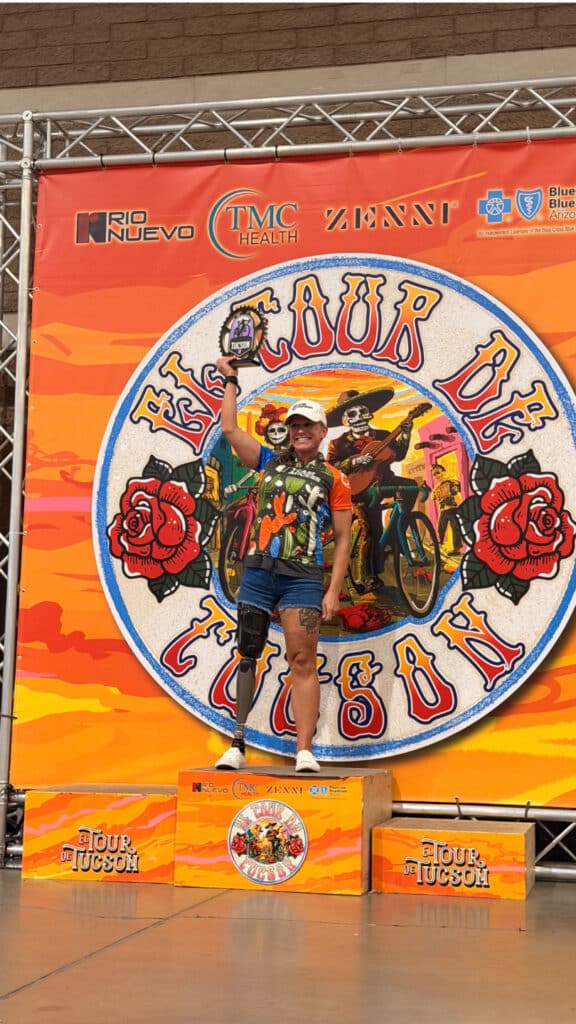 Adaptive cycling division winner celebrates on the El Tour de Tucson podium while raising a trophy above her head.