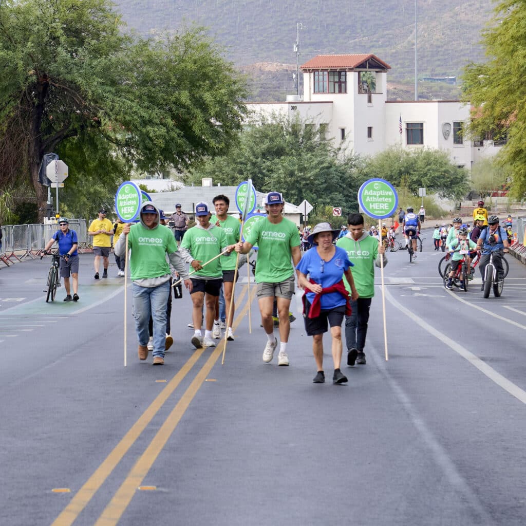 Volunteers walk along the El Tour de Tucson course holding signs that direct adaptive athletes to the event staging area.