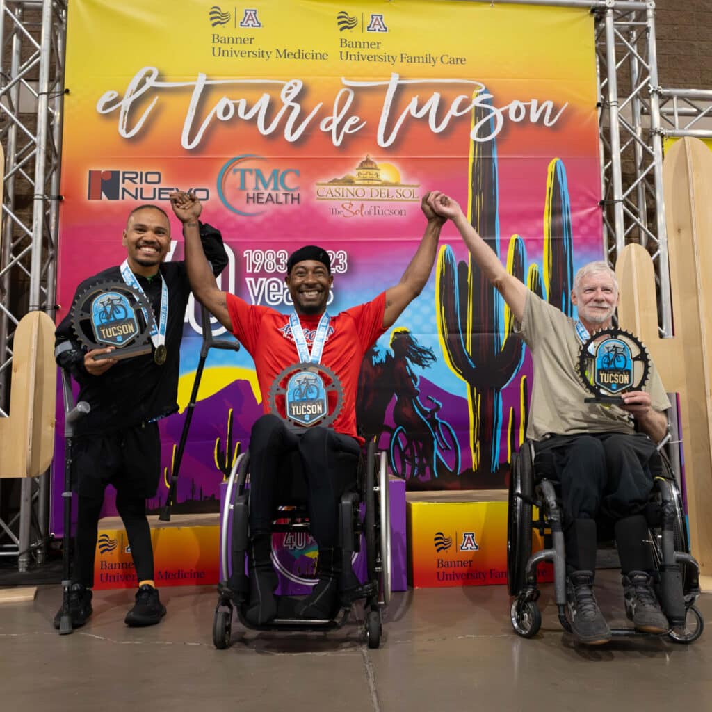 Three adaptive cycling race winners celebrate on the El Tour de Tucson awards stage. The athletes raise their joined hands in victory while holding trophy plaques and wearing medals, with an El Tour de Tucson event backdrop and sponsor logos behind them.