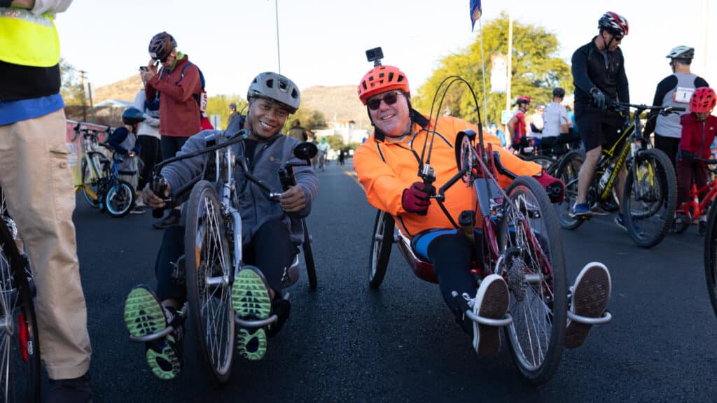 Two adaptive cyclists smile while seated on handcycles at the El Tour de Tucson starting area before the race.