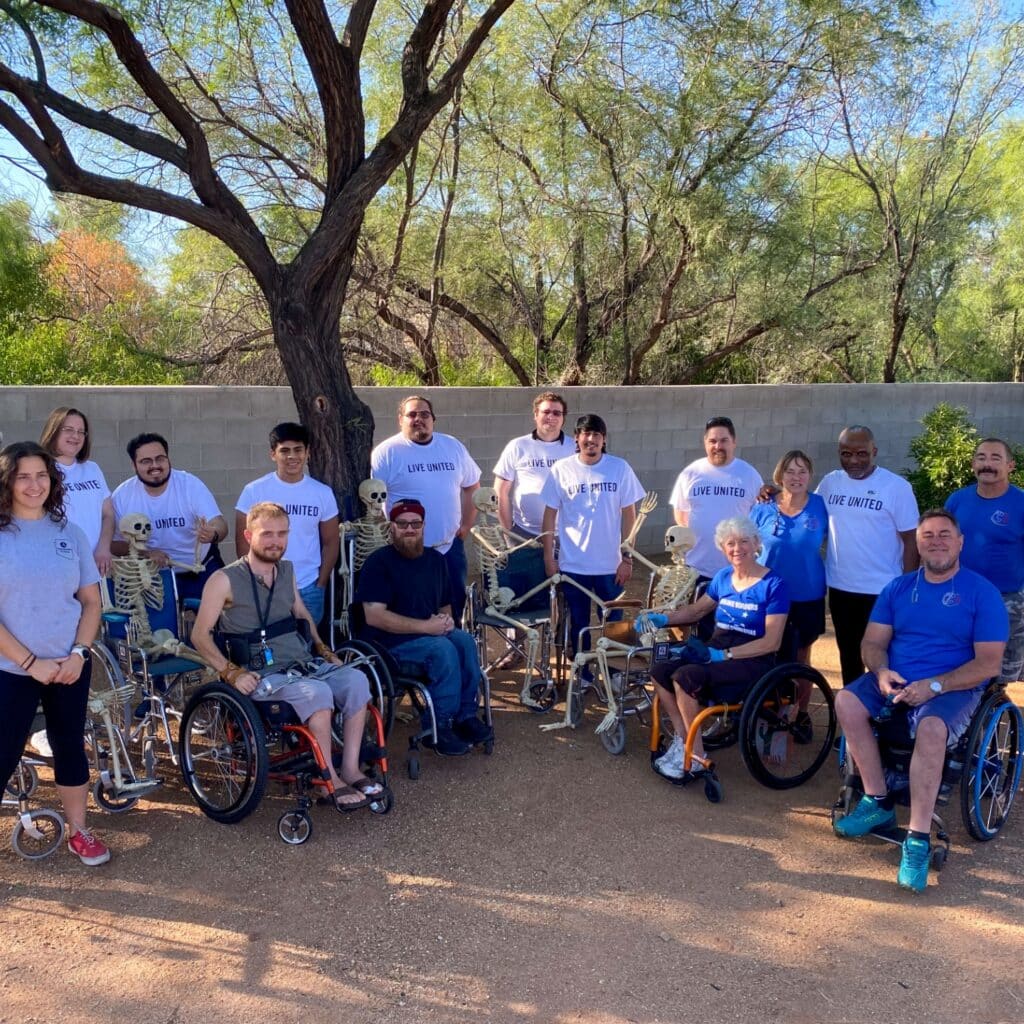 Volunteers and adaptive athletes pose together outdoors with Halloween skeleton decorations during a Southern Arizona Adaptive Sports community event.