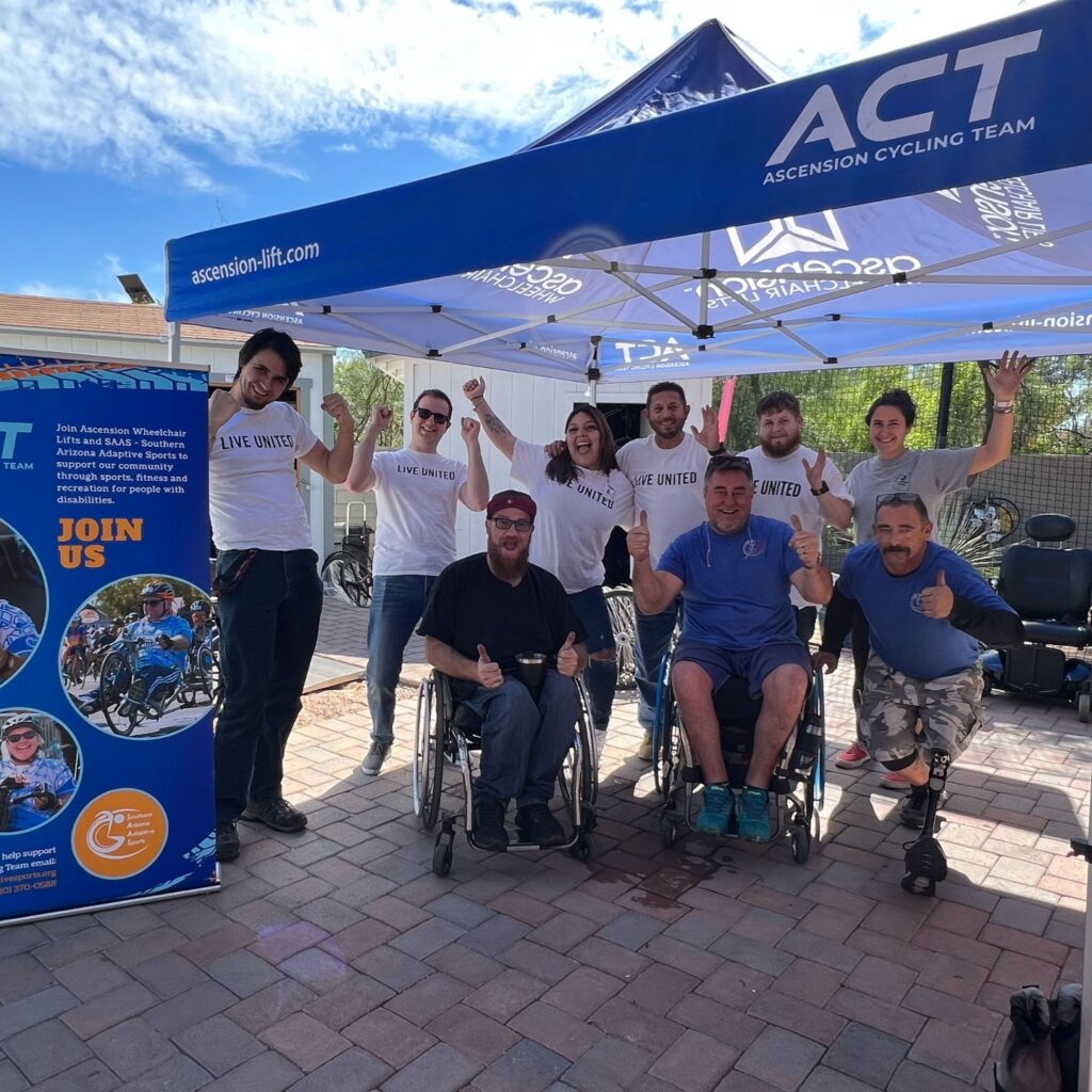 Volunteers and adaptive athletes smile together beneath an Ascension Cycling Team tent during a Southern Arizona Adaptive Sports outreach event.